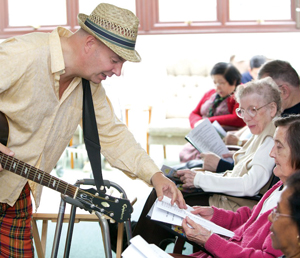 Man with a hat and guitar leaning over to turn the page of an elderly ladies music sheet